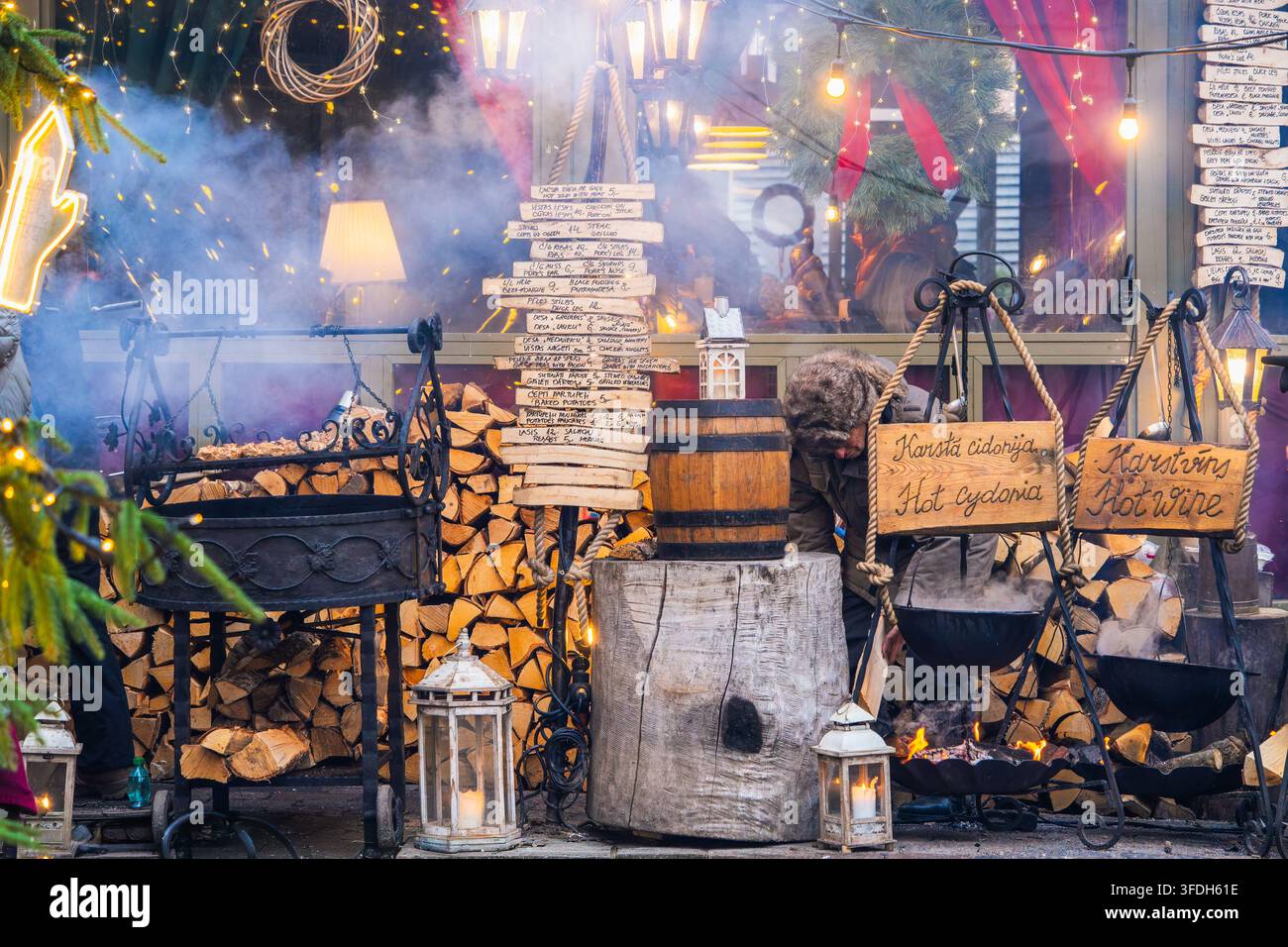 Chaudron de bois de chauffage accueillant combine des casseroles en fer suspendues, des bûches empilées et des lanternes à côté d’un foyer extérieur au marché de Noël de Riga pour des repas rustiques an Banque D'Images