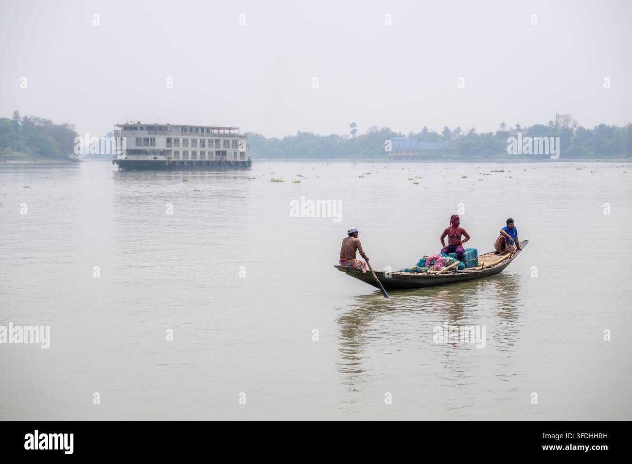Un bateau en bois traditionnel avec trois hommes rame devant un grand navire de croisière fluviale moderne au loin par une journée brumeuse sur le Gange Banque D'Images