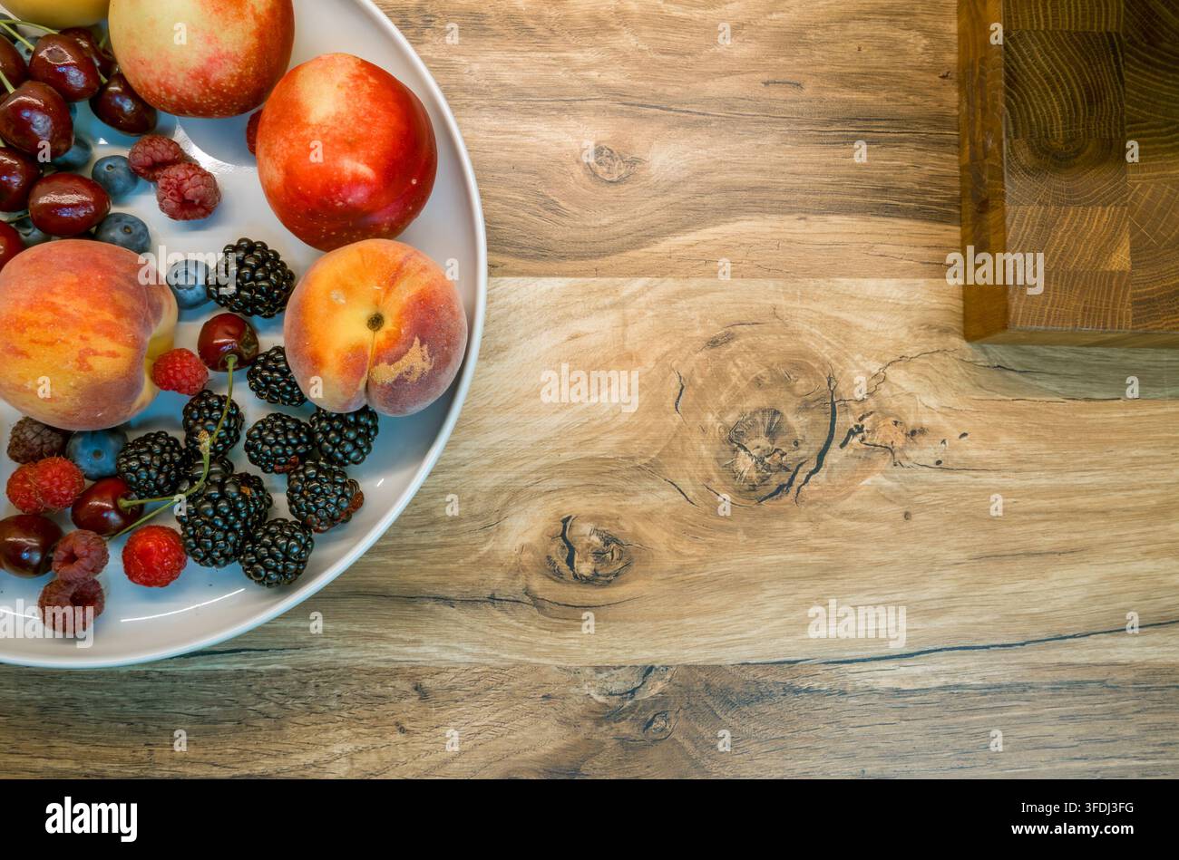 Vue aérienne de pêches, nectarines, framboises, myrtilles, cerises et mûres disposées sur une assiette blanche à côté d'une planche à découper en bois Banque D'Images