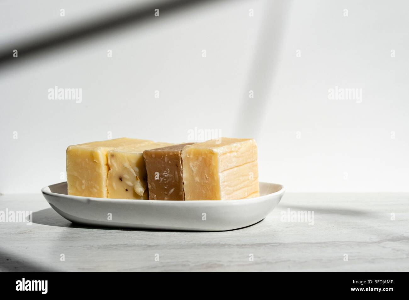 Plusieurs savons artisanaux faits main disposés sur une simple assiette en céramique blanche sous une lumière naturelle douce. Les blocs de savon rustiques présentent des variations naturelles Banque D'Images