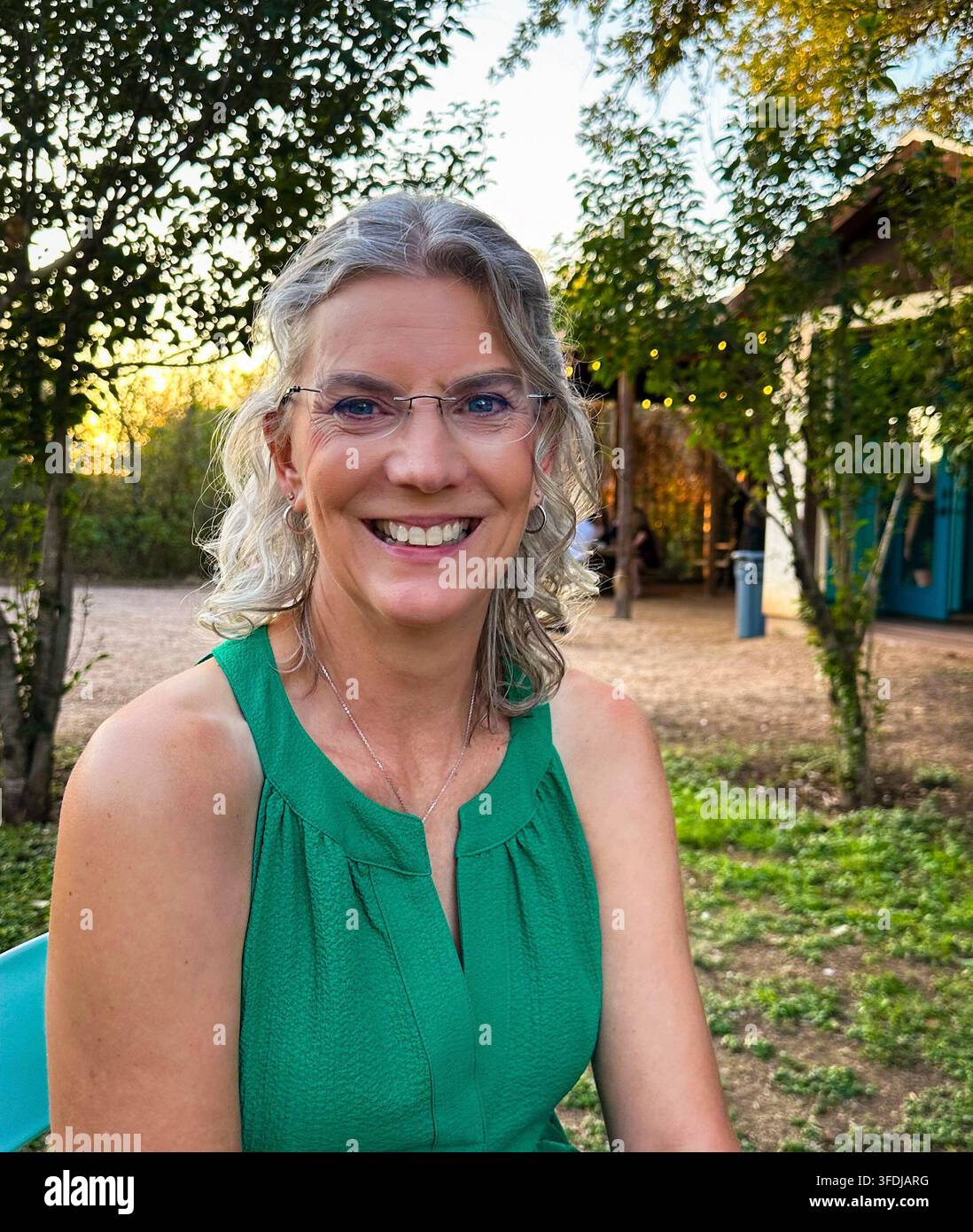 Une femme joyeuse aux cheveux gris bouclés et aux lunettes sourit chaleureusement en plein air dans un cadre de jardin à l'heure dorée, portant un haut vert avec des guirlandes lumineuses an Banque D'Images