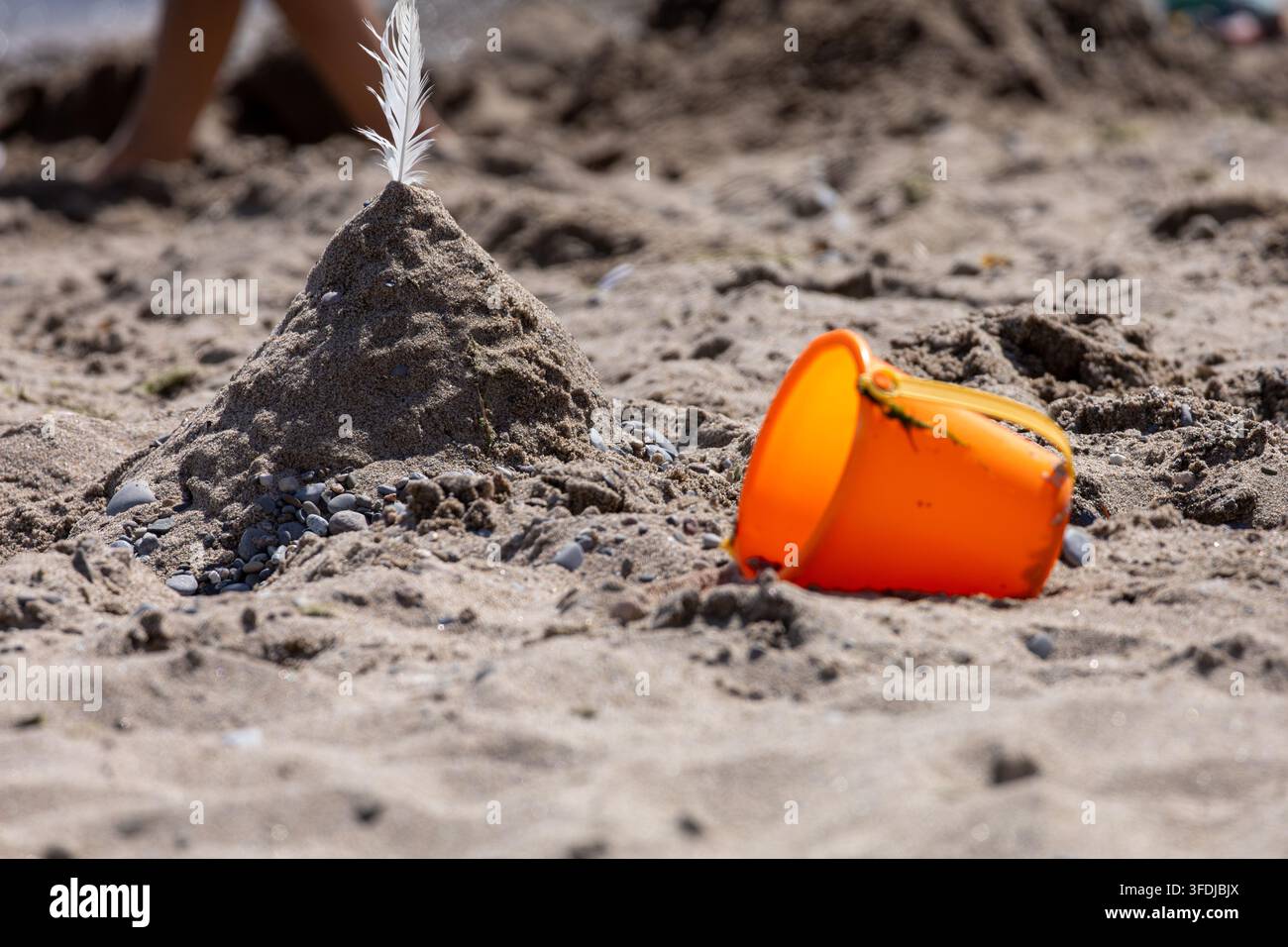 Seau de plage orange reposant à côté d'un monticule de sable sculpté à Woodbine Beach à Toronto avec une plume blanche, sous un ciel d'été lumineux. Banque D'Images