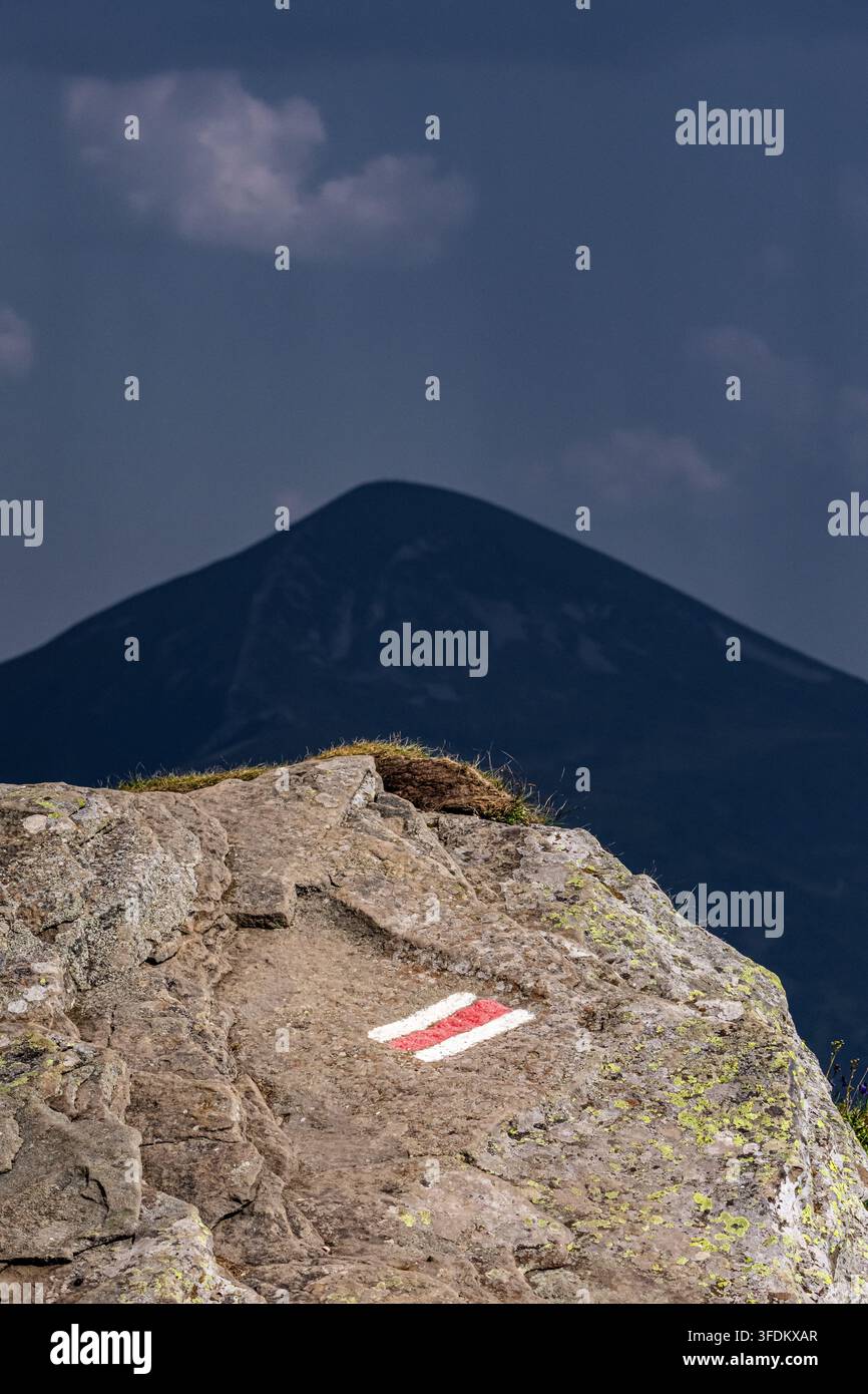 Un marqueur de piste rouge et blanc audacieux se dresse sur une pierre ensoleillée. Un pic pyramidal sombre s'élève en arrière-plan sous des nuages menaçants. Les contrastes forts transmettent di Banque D'Images