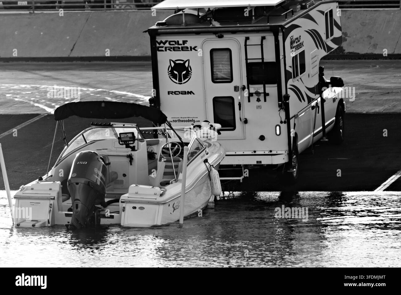 Bateau à moteur blanc avec moteur hors-bord noir flotte sur l'eau calme. Un auvent noir recouvre le cockpit. Une remorque se trouve derrière le bateau sur la rive. Teintes du coucher de soleil li Banque D'Images