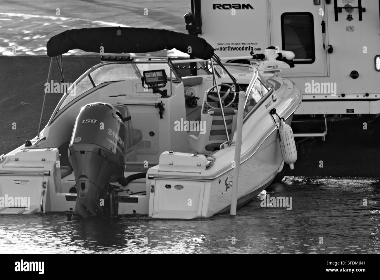 Bateau à moteur blanc avec moteur hors-bord noir flotte sur l'eau calme. Un auvent noir recouvre le cockpit. Une remorque se trouve derrière le bateau sur la rive. Teintes du coucher de soleil li Banque D'Images