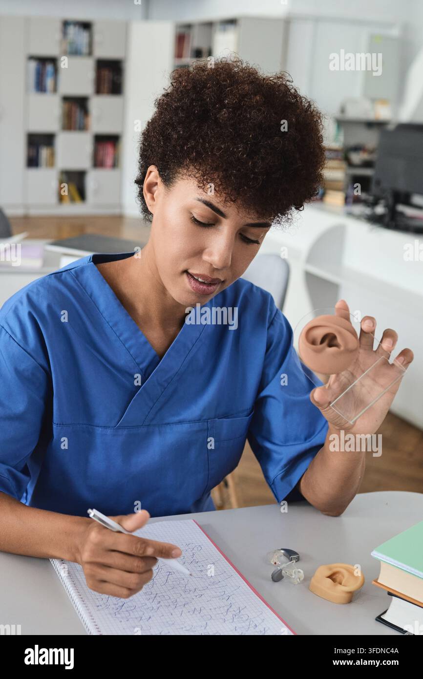 Grossissement vertical d'un étudiant en médecine afro-américain tenant un modèle anatomique d'oreille tout en prenant des notes. Audiologie clinique et diagnostic auditif Banque D'Images