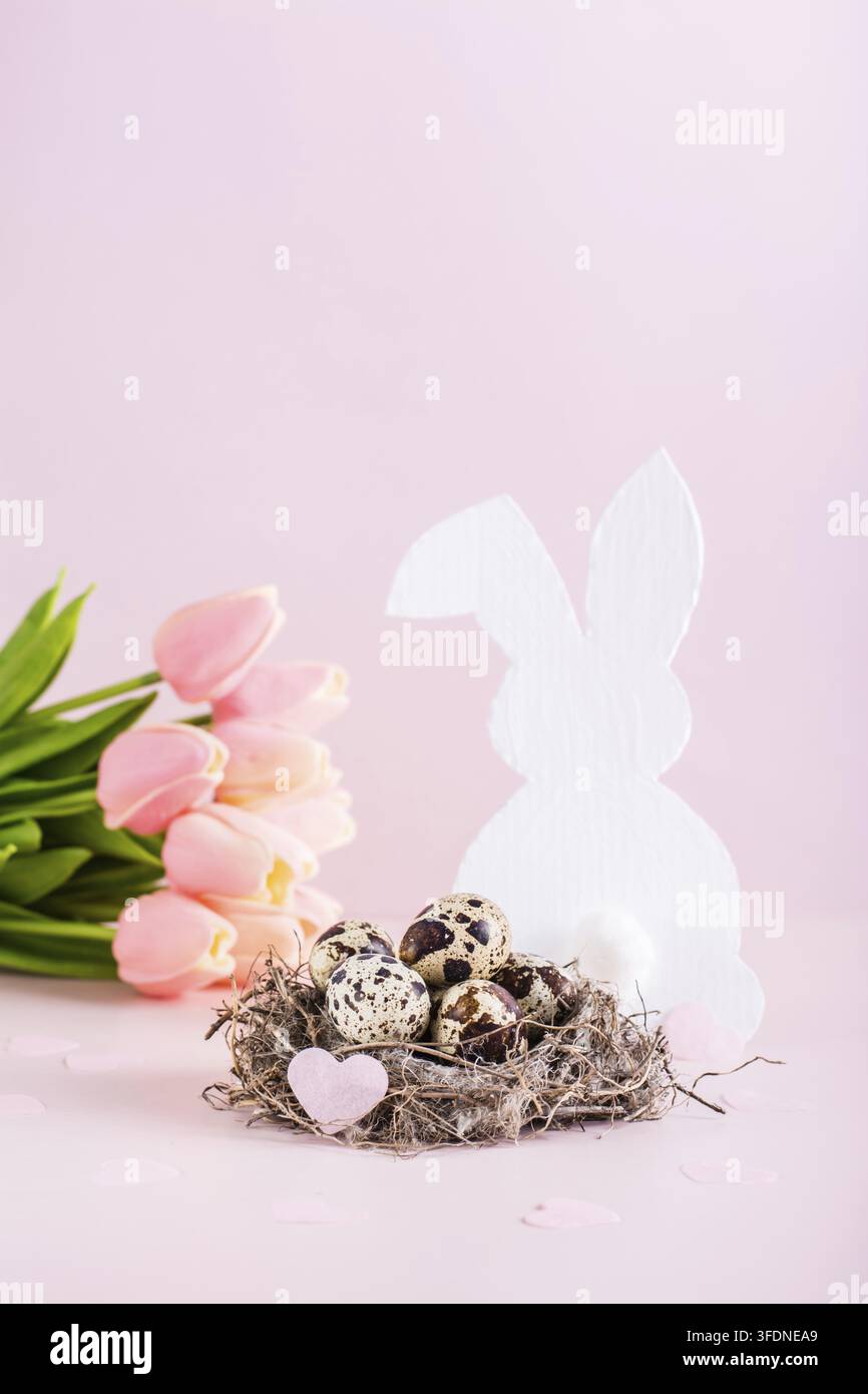 Œufs de caille de Pâques joyeux dans un nid, lapin blanc et tulipes sur un fond rose, vue verticale Banque D'Images
