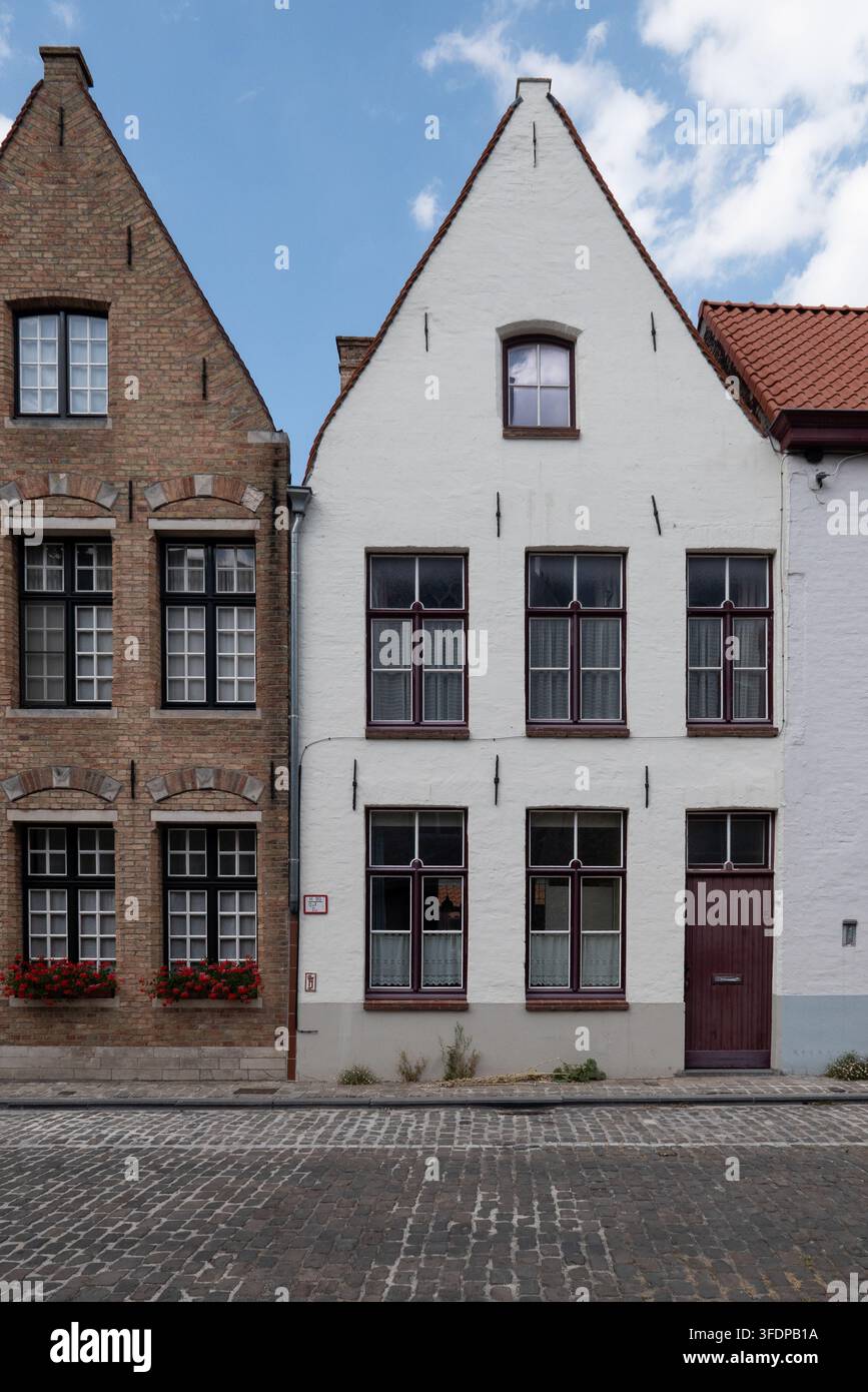 Des maisons traditionnelles en briques et à pignons blancs avec de hautes fenêtres et des fleurs rouges bordent une rue pavée dans le centre historique de la ville. Bruges, Belgique Banque D'Images