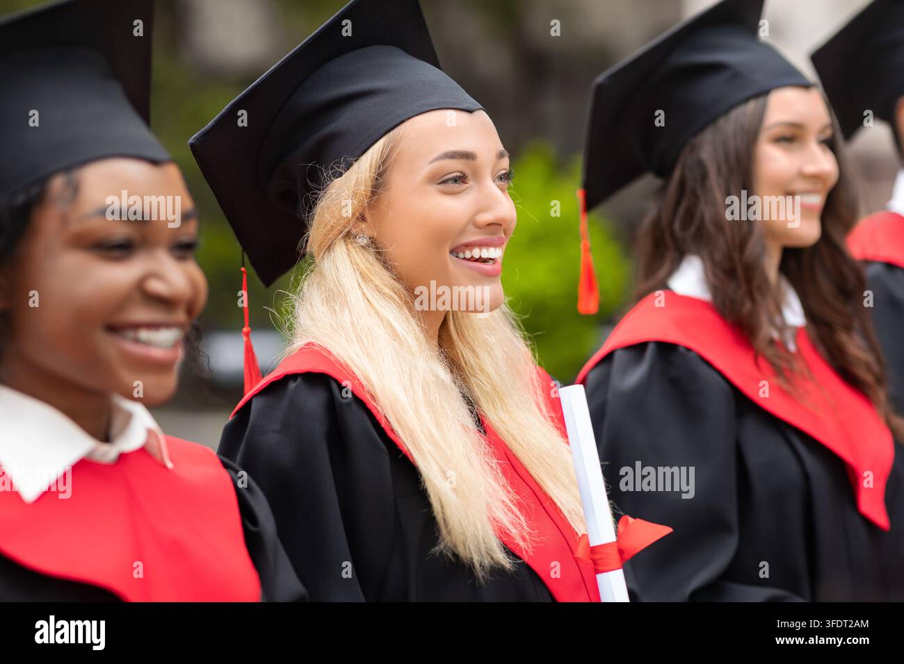 Jeune femme blonde et jolie souriante au milieu d'un groupe international d'étudiants, portant des costumes de graduation et tenant des diplômes, diplômée multiraciale Banque D'Images