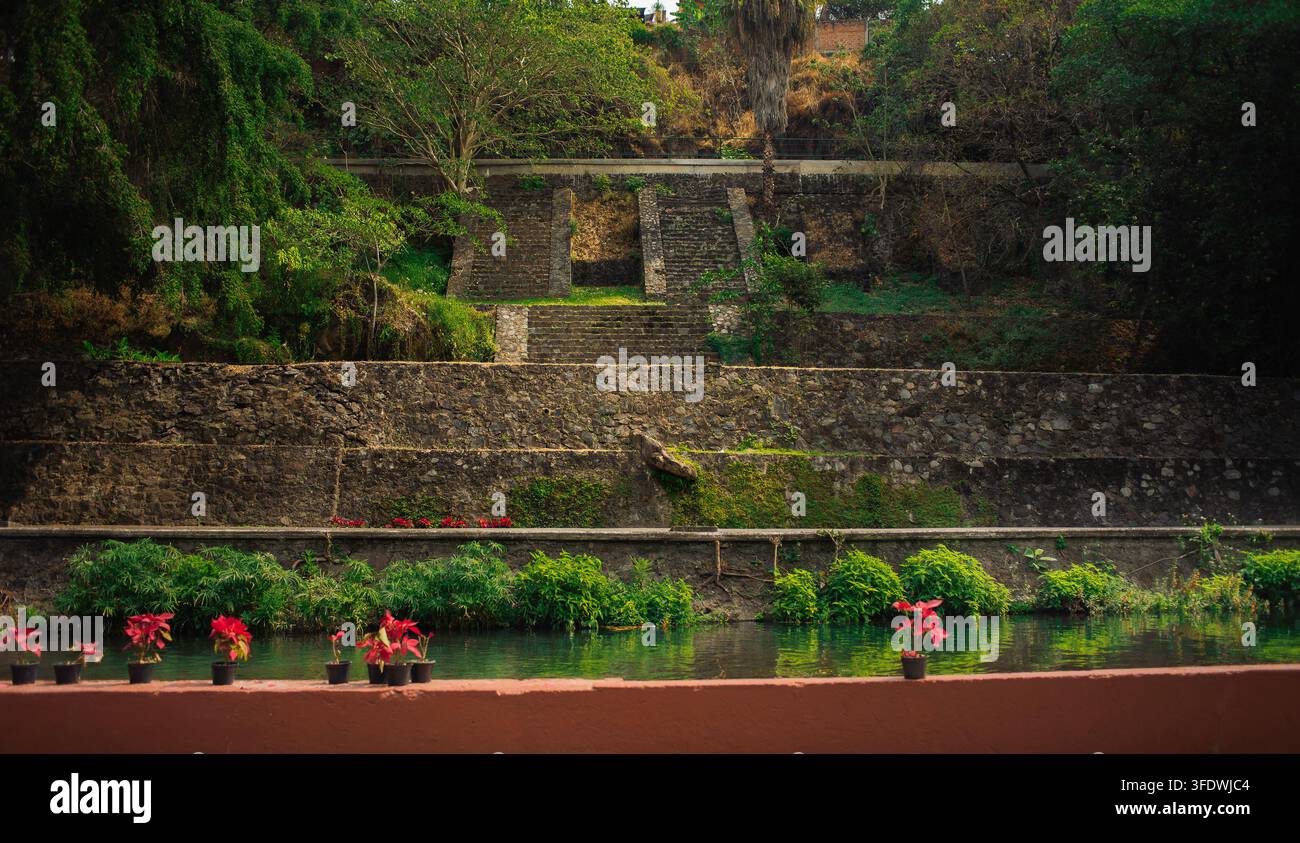 Cuernavaca, Morelos, 03 03 2026. Structure pyramidale ancienne se dresse à côté d'une rivière calme entourée de plantes de poinsettia rouges vives à l'intérieur de Chapultepec P Banque D'Images