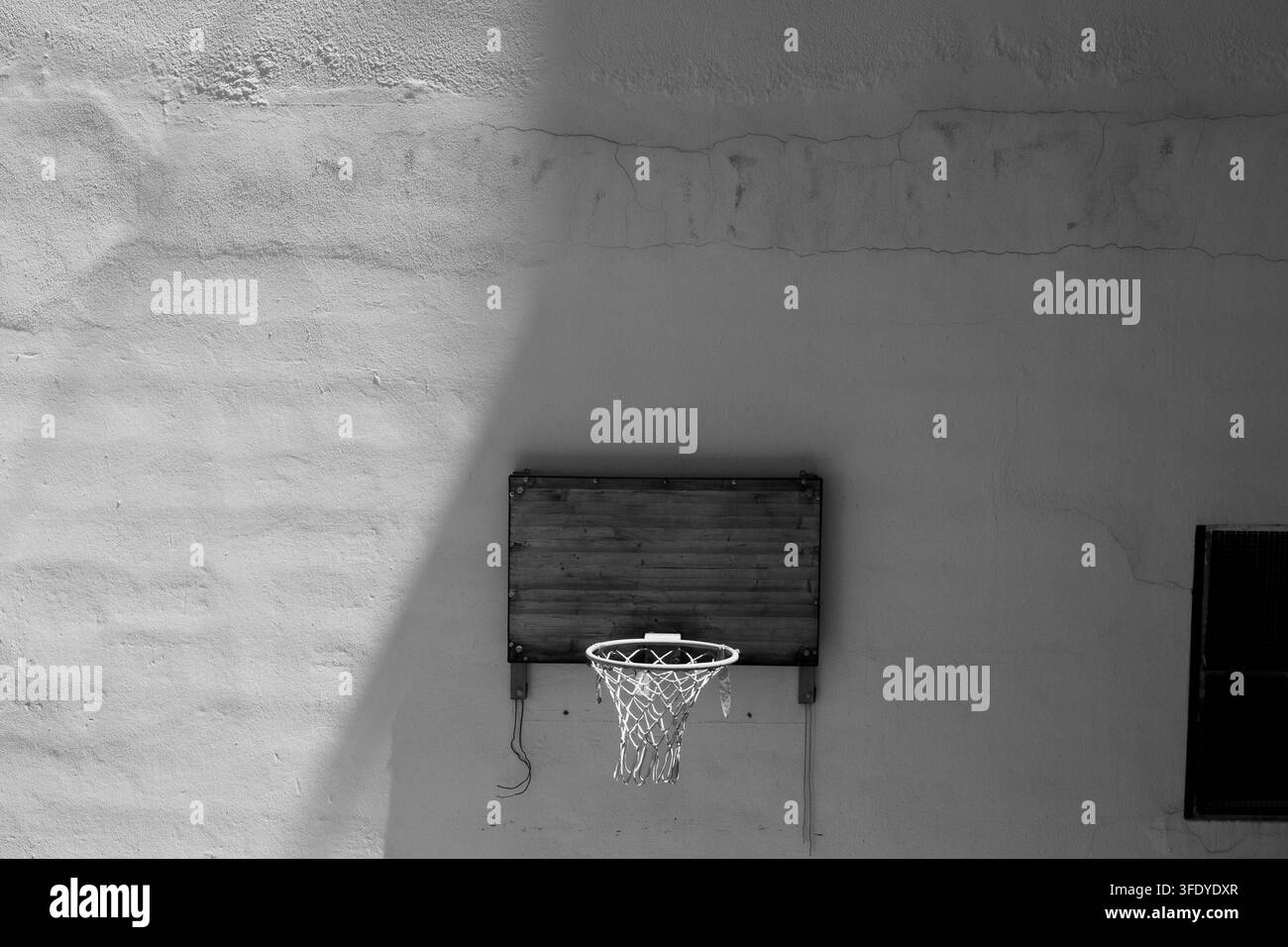 Un cliché minimaliste en noir et blanc d'un panier de basket sur un mur texturé. Des ombres diagonales dures créent une atmosphère urbaine de contraste élevé et sombre. Banque D'Images