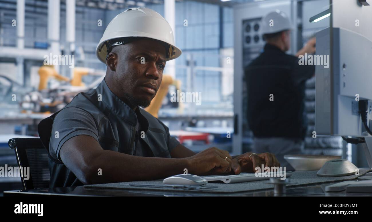 Dans le bureau de contrôle automatisé des plantes, un spécialiste afro-américain tape sur un clavier PC pour gérer les algorithmes de production. Les techniciens réseau entretiennent le matériel des serveurs en arrière-plan. Industrie 4.0. Banque D'Images