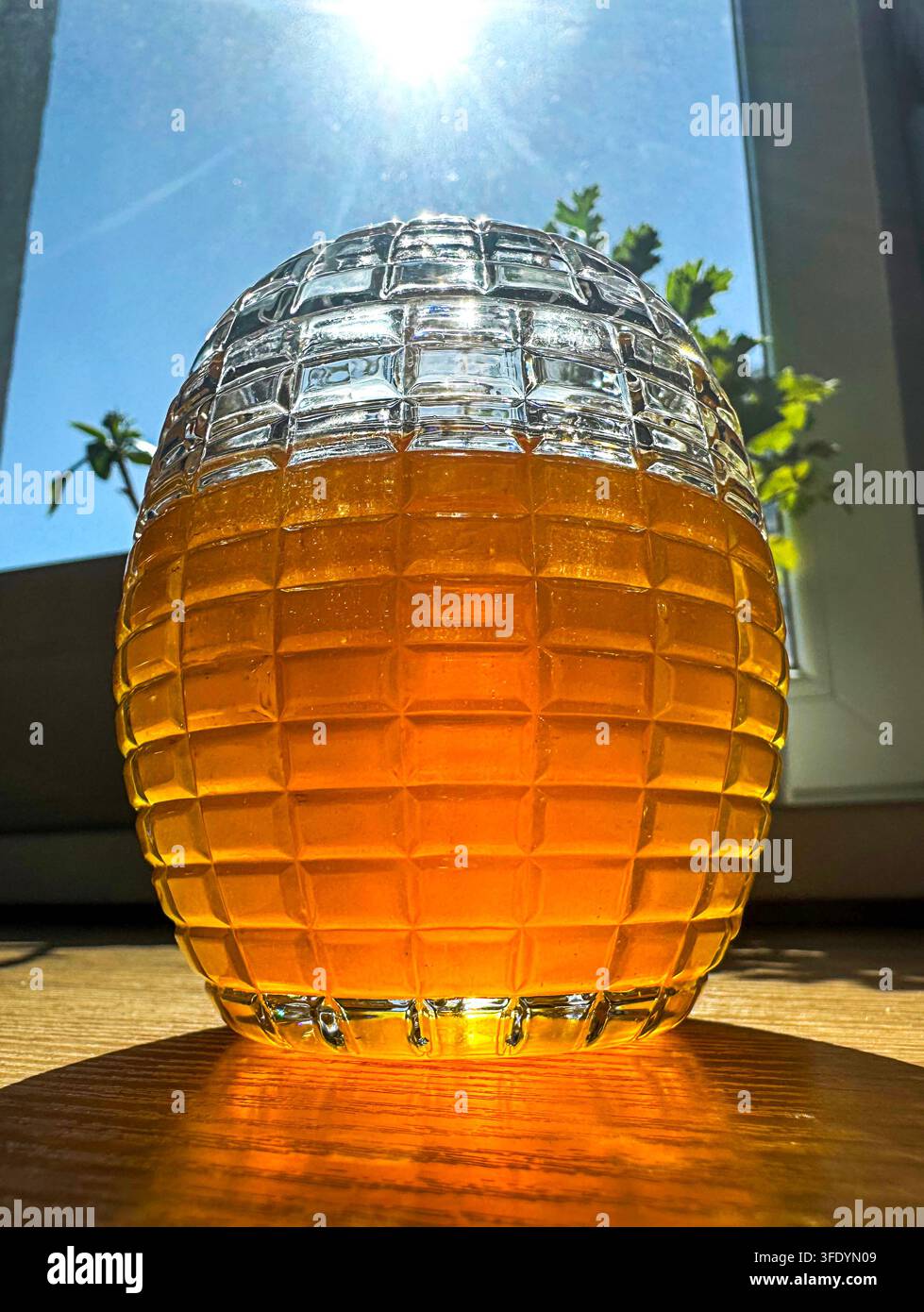 Pot de miel en verre texturé brillant d'une couleur ambrée à la lumière du soleil sur un rebord de fenêtre en bois Banque D'Images
