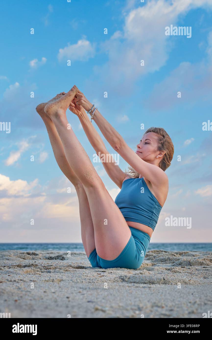 Une femme pratique l'Ubhaya Padangusthasana yoga, Both Big Toe Pose, en équilibre assis sur le sable de la plage, tenant ses deux pieds levés sous un ciel bleu Banque D'Images