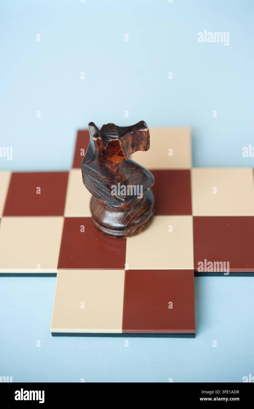 Gros plan d'un chevalier en bois sombre debout sur une petite croix d'échecs rouge et crème devant un fond bleu pâle Banque D'Images