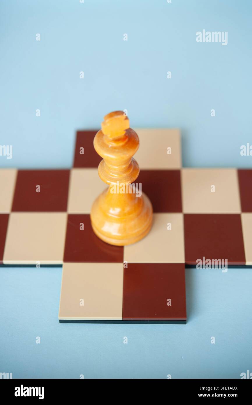 Gros plan d'un roi d'échecs en bois clair posé sur une petite croix d'échecs rouge et crème sur un fond bleu pâle Banque D'Images
