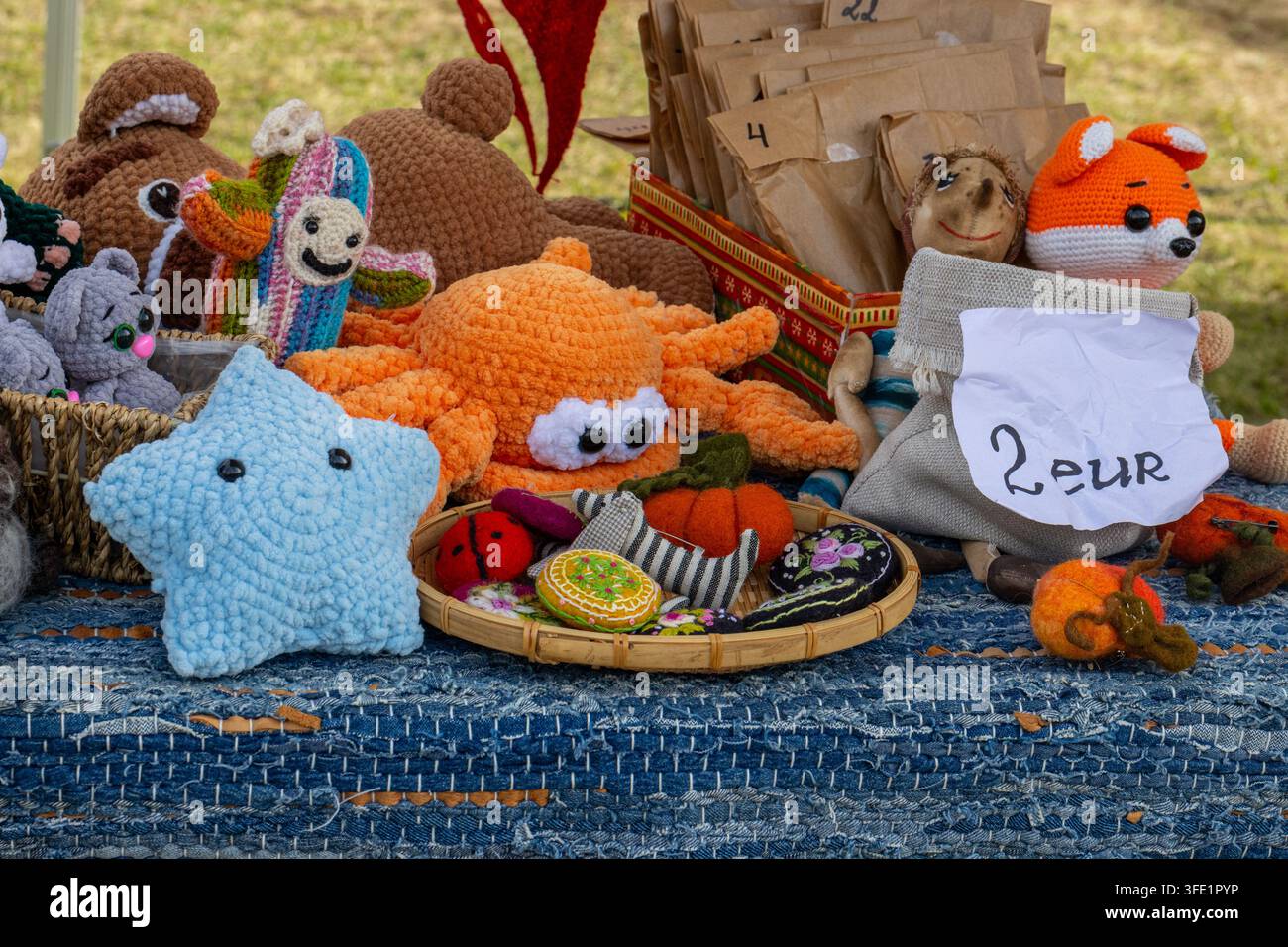 Collection colorée de jouets en peluche amigurumi crochetés à la main et d'accessoires feutrés disposés sur une nappe bleue tissée à un marché artisanal en plein air Banque D'Images