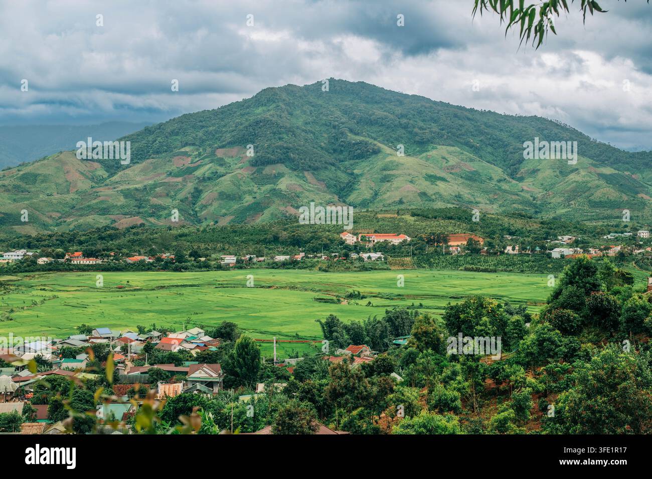 Montagne verdoyante s'élevant au-dessus des champs agricoles et d'une colonie rurale sous un ciel nuageux Banque D'Images