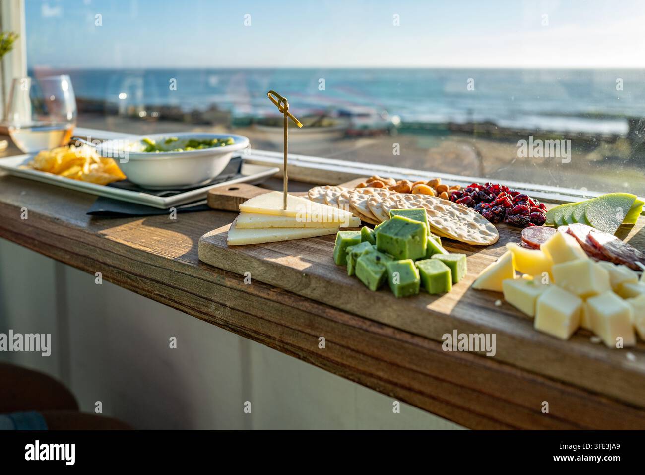 Planche de charcuterie gourmet avec divers fromages et crackers servis sur la rambarde d'un balcon donnant sur le littoral de l'océan Pacifique. Banque D'Images