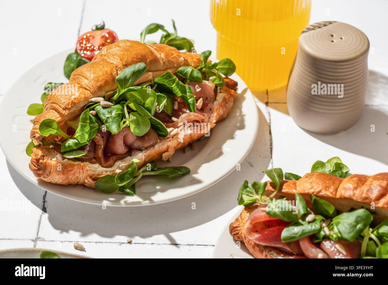Sandwich croissant avec prosciutto, fromage frais, salade de maïs et tomates cerises sur une assiette blanche posée sur une table en carrelage blanc, servi pour le petit-déjeuner matinal Banque D'Images
