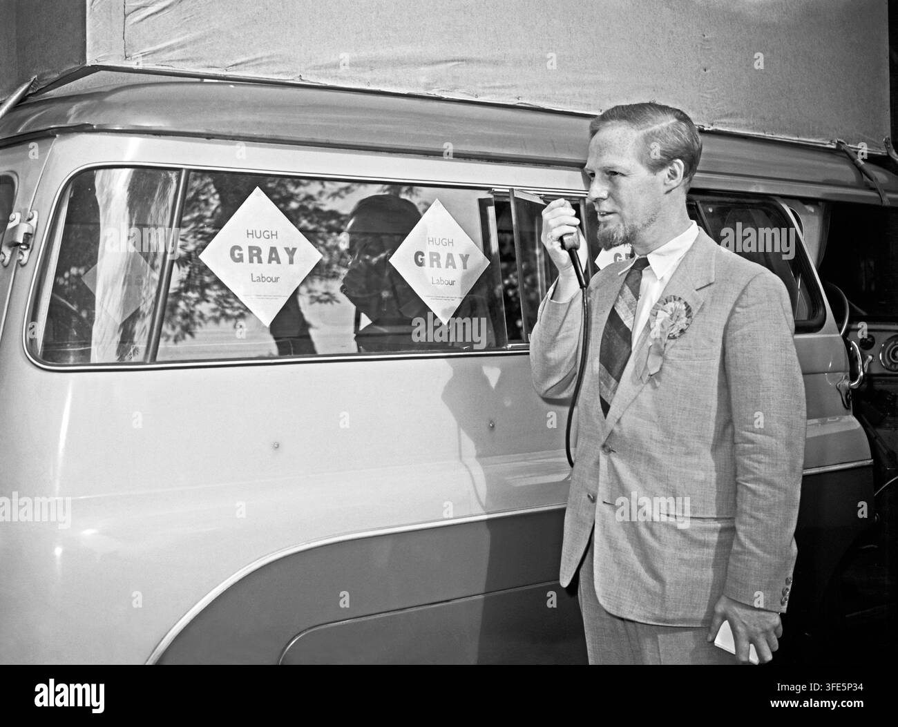 Candidat parlementaire du Parti travailliste, Hugh Gray, en campagne de porte-à-porte lors des élections générales de 1959 dans la circonscription d'Aylesbury, Buckinghamshire, Angleterre, Royaume-Uni. Ici, il parle dans un microphone diffusant ses mots par des haut-parleurs cachés sous une bâche, montés au sommet de son minibus ou bus électoral. Il porte la rosette du parti. M. Gray est arrivé deuxième derrière le candidat conservateur réélu Spencer Summers. Banque D'Images