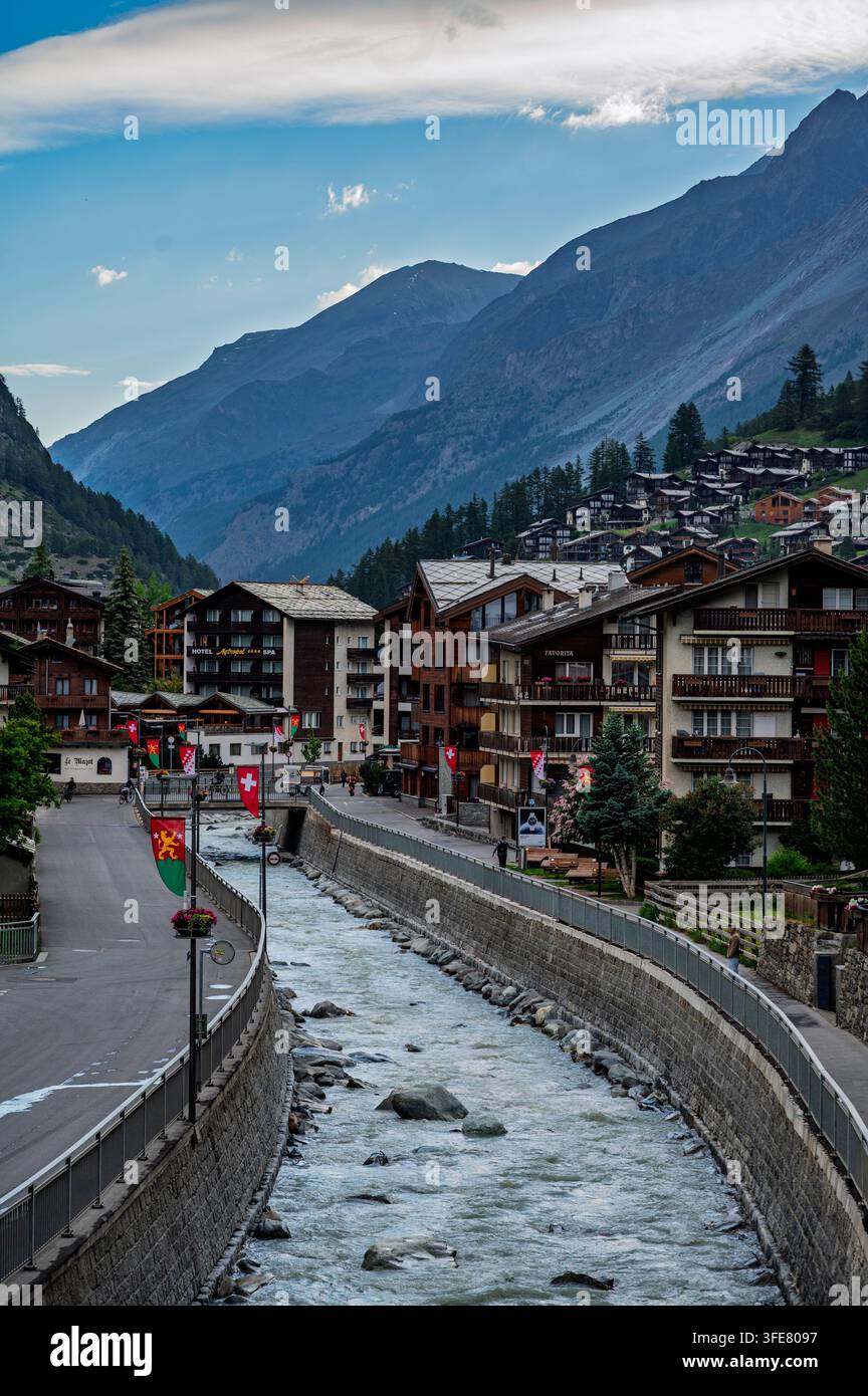 Zermatt, Suisse - Juin 2026 : Un ruisseau de montagne impétueux traverse le village de Zermatt, bordé de chalets en bois, de panneaux de magasins et de f héraldique Banque D'Images