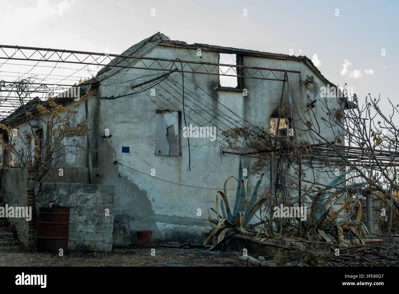 Une maison rurale abandonnée avec du plâtre fissuré, des fenêtres brisées, des fils emmêlés et une végétation sèche envahissante se dresse sous un ciel lumineux. Banque D'Images