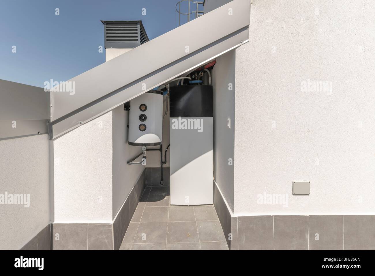 Coin utilitaire sur le toit avec chauffe-eau, tuyaux apparents, conduit de ventilation et sol carrelé gris. Banque D'Images