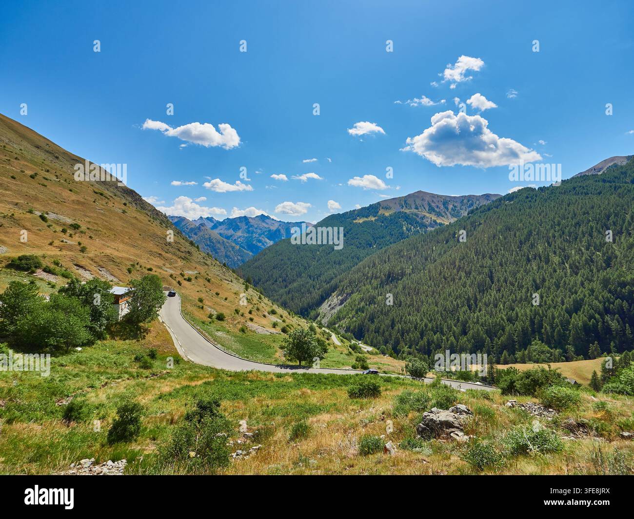 Route de montagne sinueuse à travers une vallée verte et une forêt de pins sous un ciel bleu parsemé de gros nuages Banque D'Images