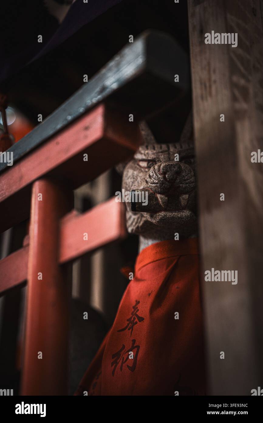 Une statue de renard en pierre usée portant un bavoir rouge avec des caractères japonais, partiellement éclairée à côté d'une structure de porte torii rouge à un sanctuaire Inari. Banque D'Images