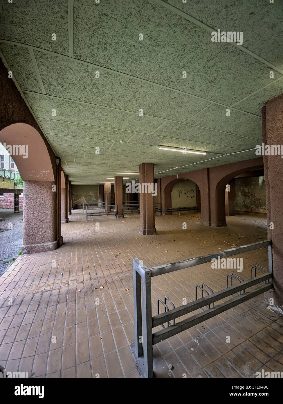 Cour abritée vide sous les arches du bâtiment, avec sol carrelé, colonnes carrées, lumières fluorescentes, murs érodés et débris éparpillés, cre Banque D'Images