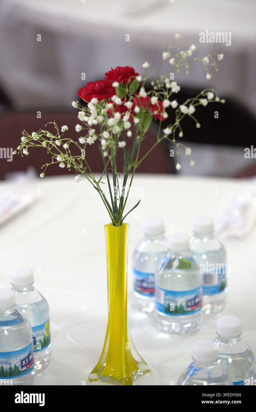 Un arrangement floral de œillets rouges et de gypsophile blanc dans un vase jaune élancé est posé sur une table entourée de bouteilles d'eau lors d'un événement. Banque D'Images