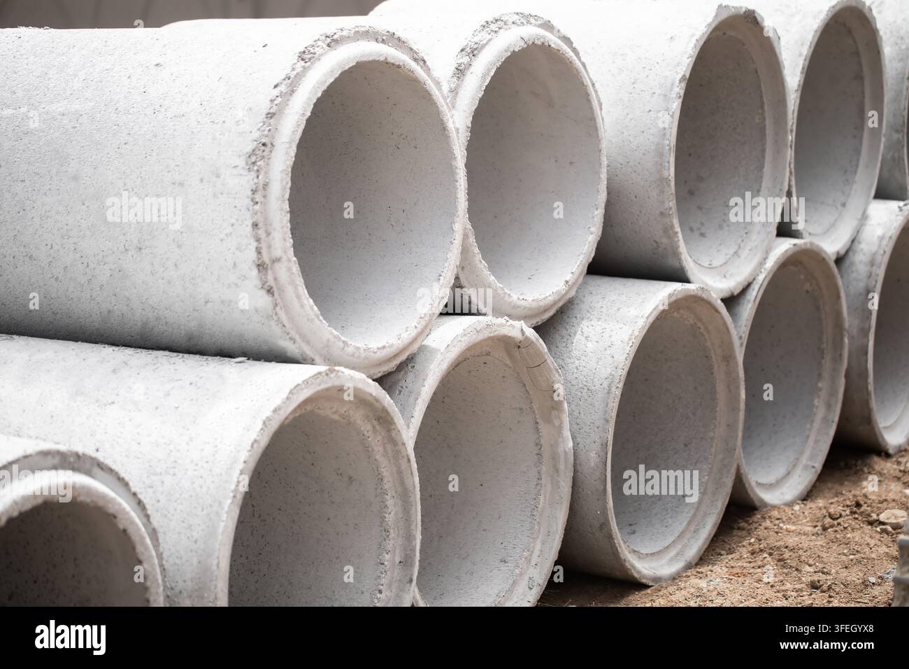 Tubes de drainage en béton empilés en formation pyramidale sur un chantier de construction en terre, les joints arrondis visibles le long des rangées, la lumière du matin projette des ombres douces à travers Banque D'Images