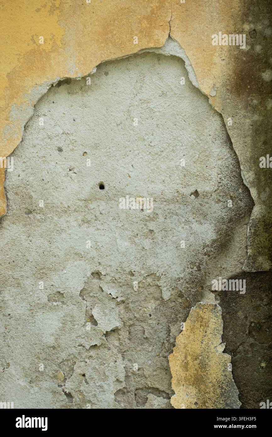 Le plâtre jaune qui s'écaille révèle un ciment gris rugueux en dessous, créant une texture de mur vieillie et abîmée avec des fissures, des taches et de la dégradation Banque D'Images