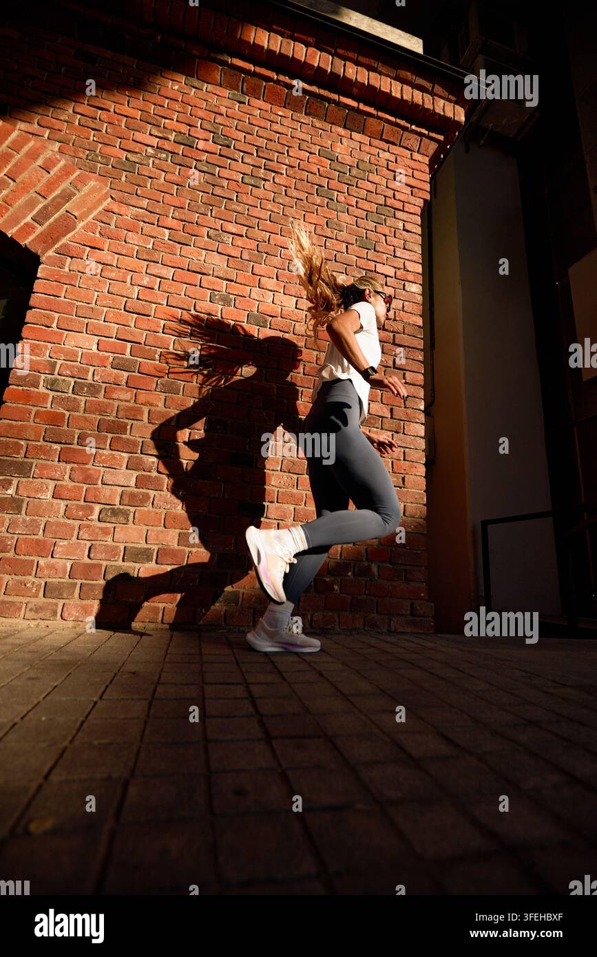Prise de vue dynamique en contre-plongée d'une femme en forme jogging sur un trottoir urbain. Ses longs cheveux blonds volent en mouvement. Une lumière solaire forte projette une ombre dramatique sur le brique rouge Banque D'Images