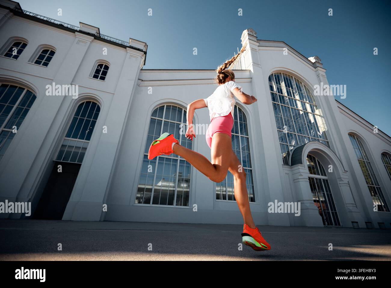 Prise de vue en contre-plongé d'une jeune femme en forme courant en plein air. Athlète en short rose et baskets orange s'entraîne en ville devant un bâtiment blanc et un ciel bleu Banque D'Images