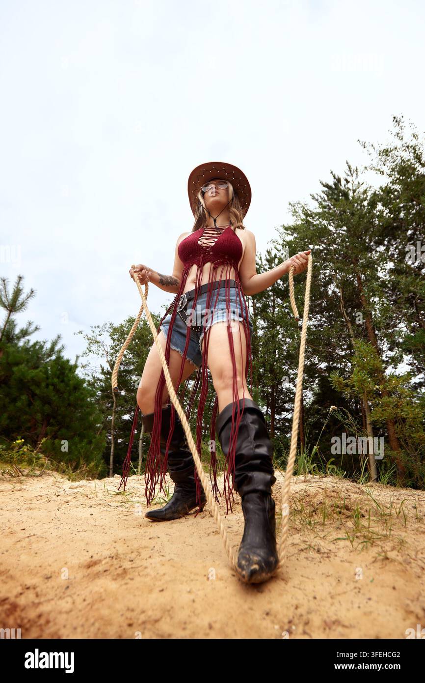 Prise de vue en bas de corps d'une jeune femme puissante de style western moderne. Elle porte un chapeau de cowboy, un haut au crochet et des bottes, tenant une corde dans un terrain sablonneux et boisé Banque D'Images