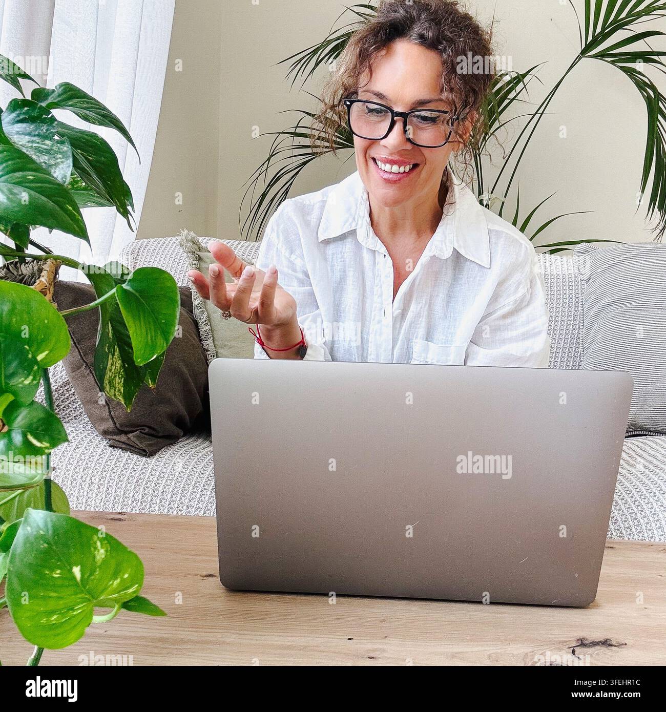 Femme portant des lunettes et une chemise blanche souriant brillamment tout en ayant un appel vidéo sur son ordinateur portable, travaillant à distance depuis son confortable bureau à domicile Banque D'Images