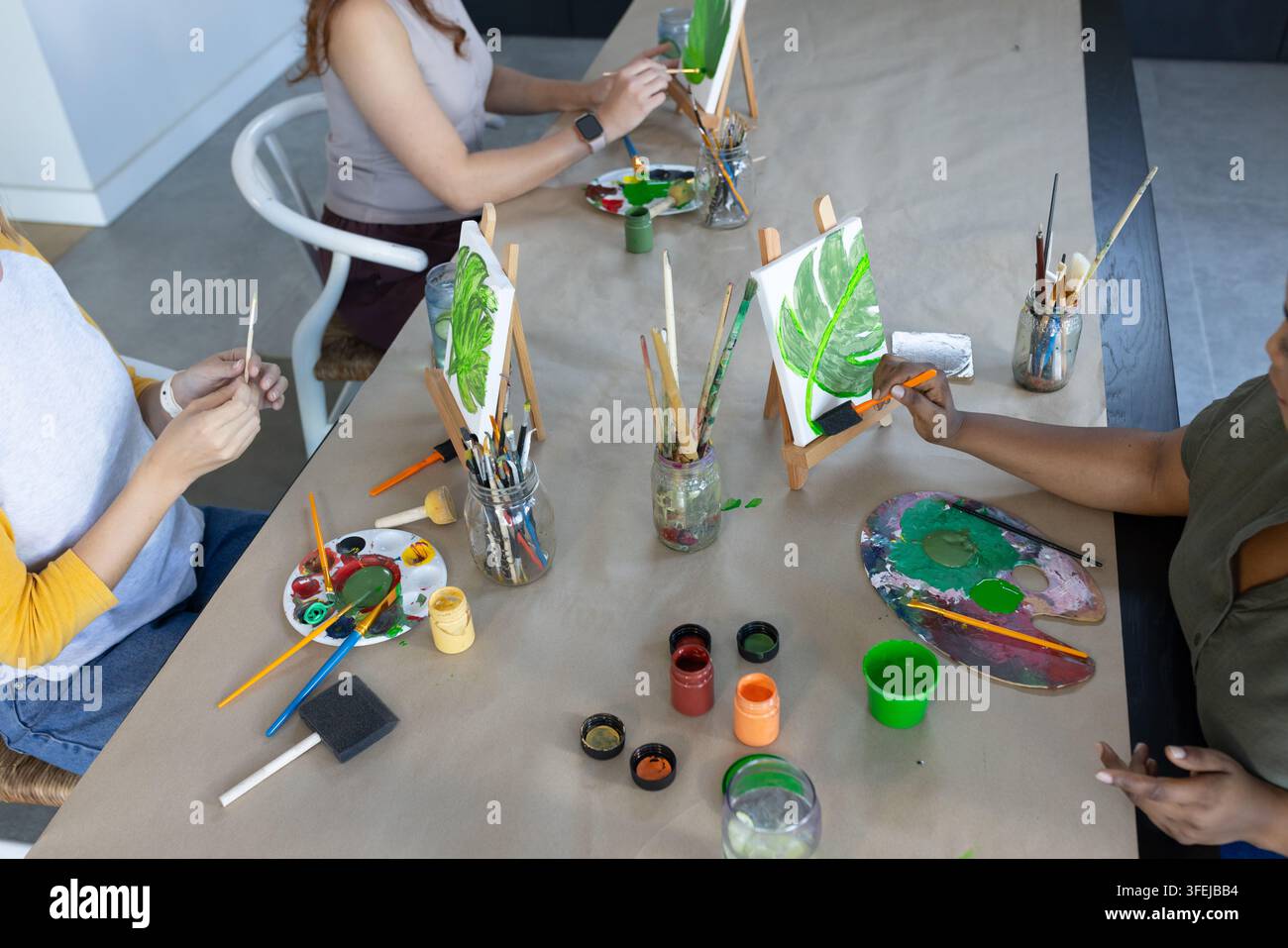 Diverses amies peignant des toiles de feuilles vertes à une table commune avec des pinceaux et des palettes Banque D'Images