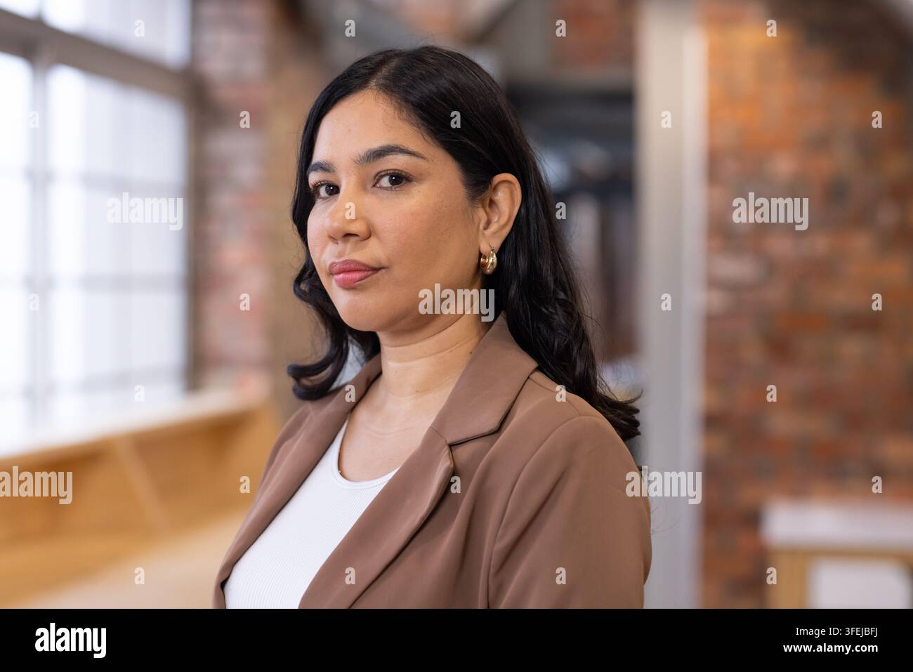 Femme professionnelle posant dans un espace de travail moderne près d'une fenêtre à multiples vitres, portant un blazer marron et des créoles en or Banque D'Images