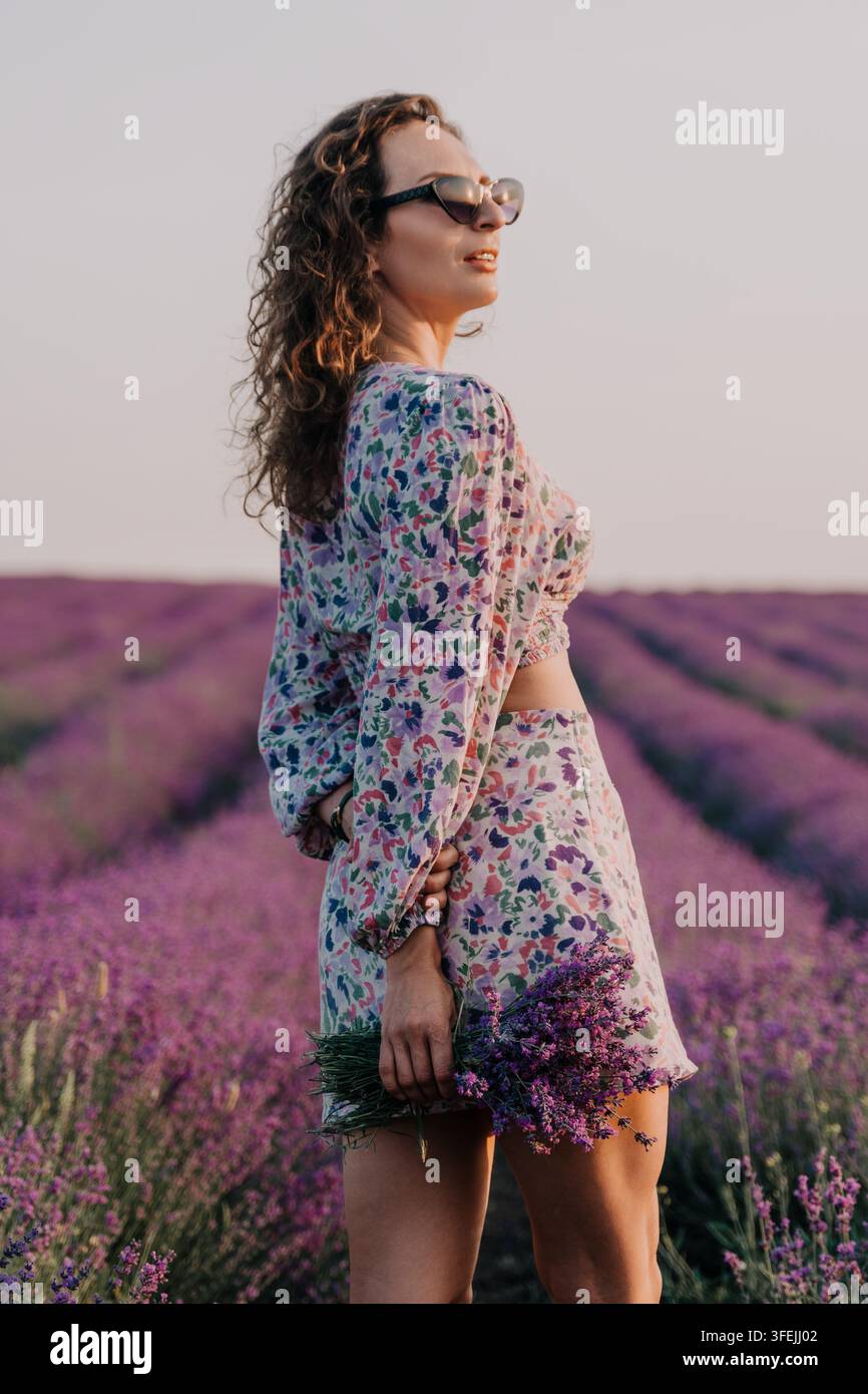 Une jeune femme portant une tenue deux pièces fleurie et des lunettes de soleil se tient dans un champ de lavande violet vif au coucher du soleil, tenant un bouquet de tiges de lavande. Banque D'Images