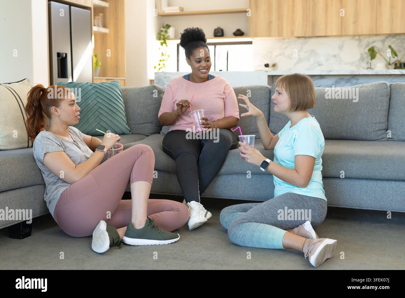 Diverses amies féminines en conversation, vêtues de vêtements de sport, sirotant des gobelets transparents avec des pailles dans un appartement Banque D'Images