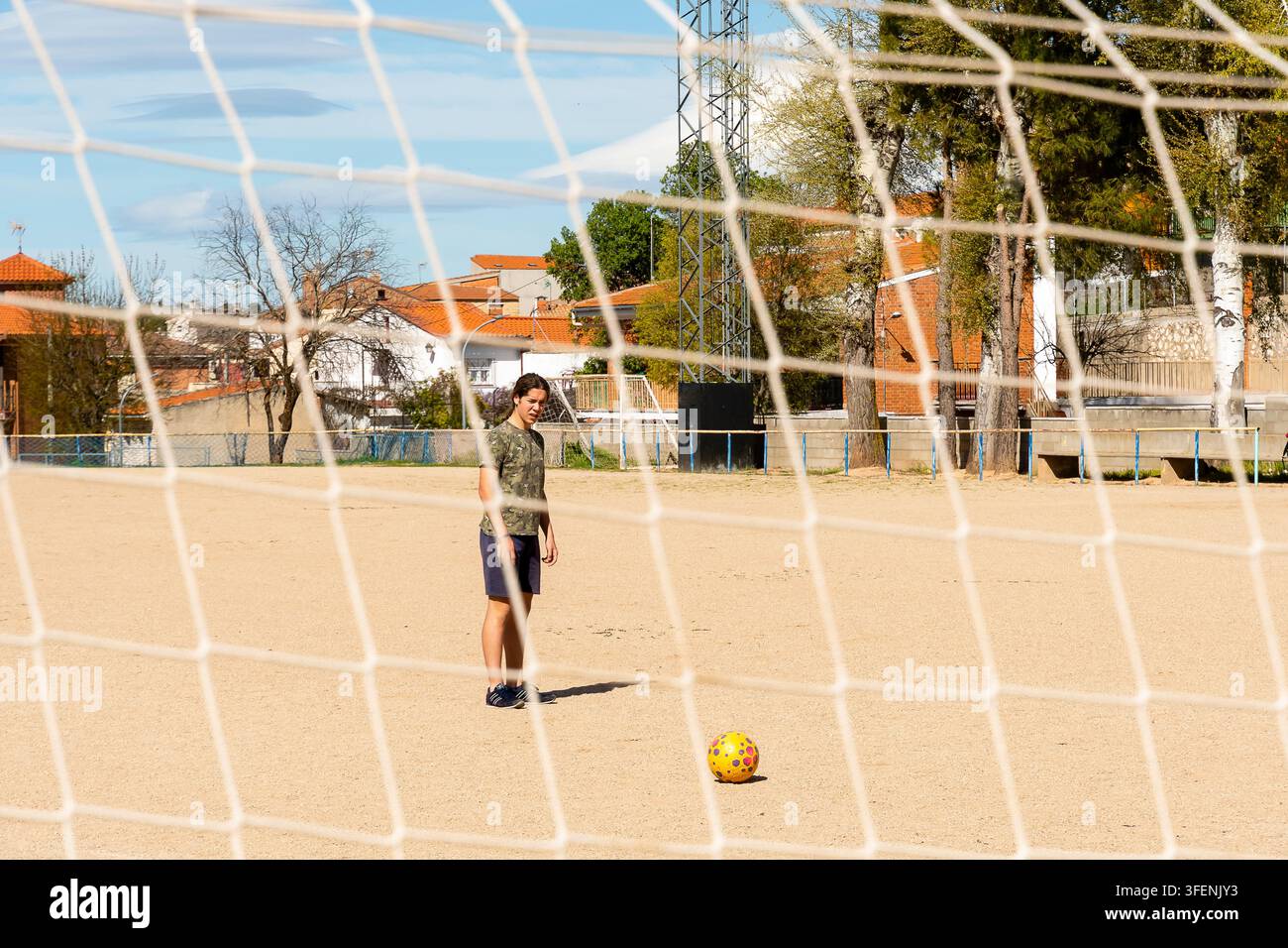 Un garçon donne un coup de pied à un ballon de football tout en se tenant sur un terrain sablonneux entouré de maisons par une journée claire. Banque D'Images