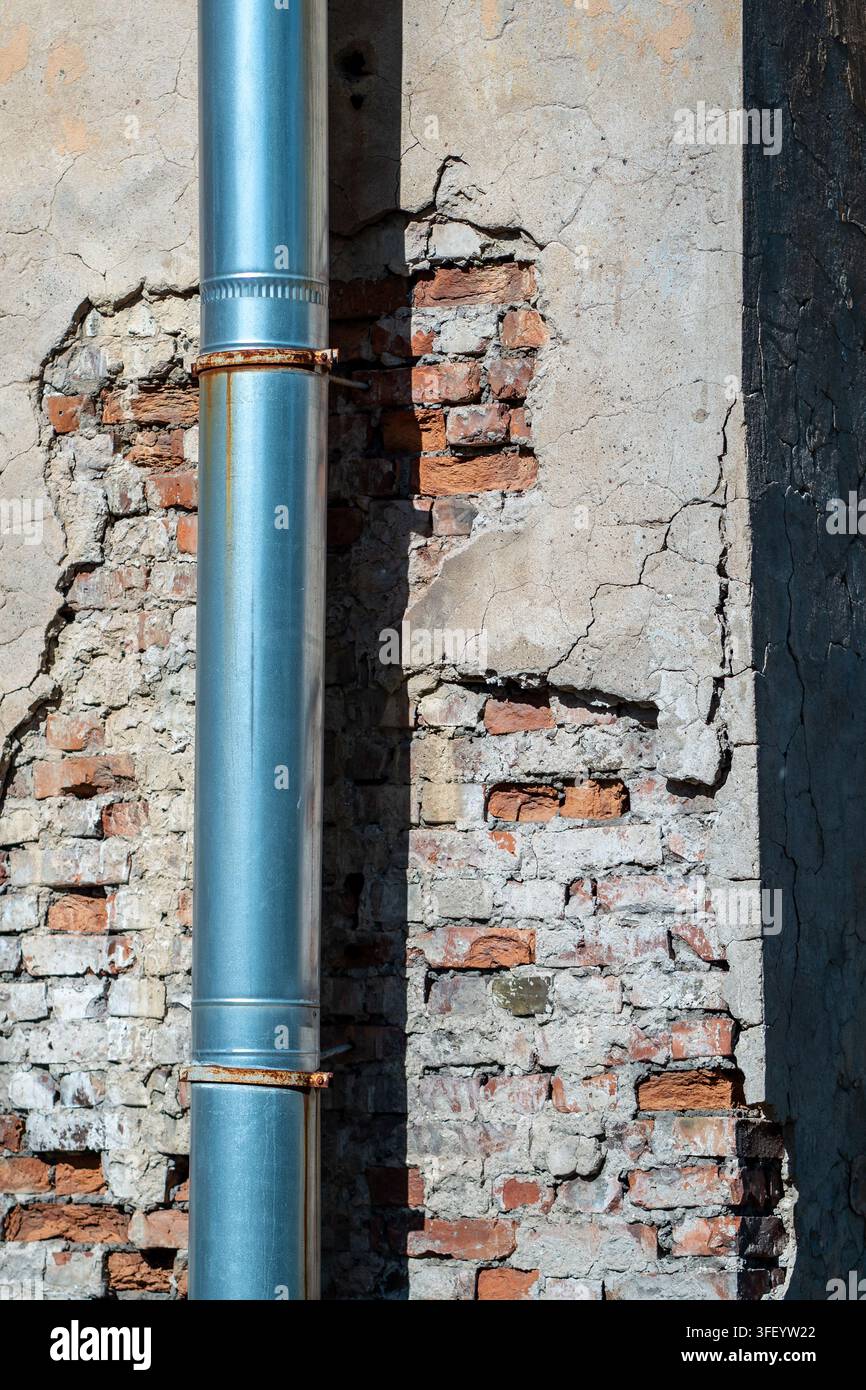 Tuyau de drainage en métal sur un vieux mur en plâtre fissuré avec brique rouge apparente, altération et rouille. Banque D'Images