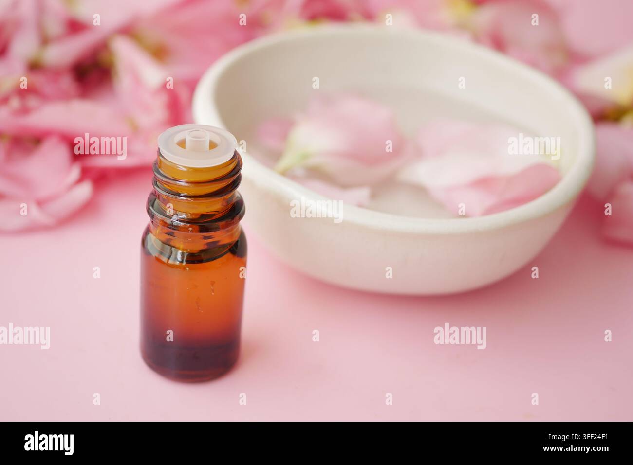 petite bouteille en verre ambrée d'huile essentielle à côté d'un bol rempli d'eau et de pétales de rose roses Banque D'Images