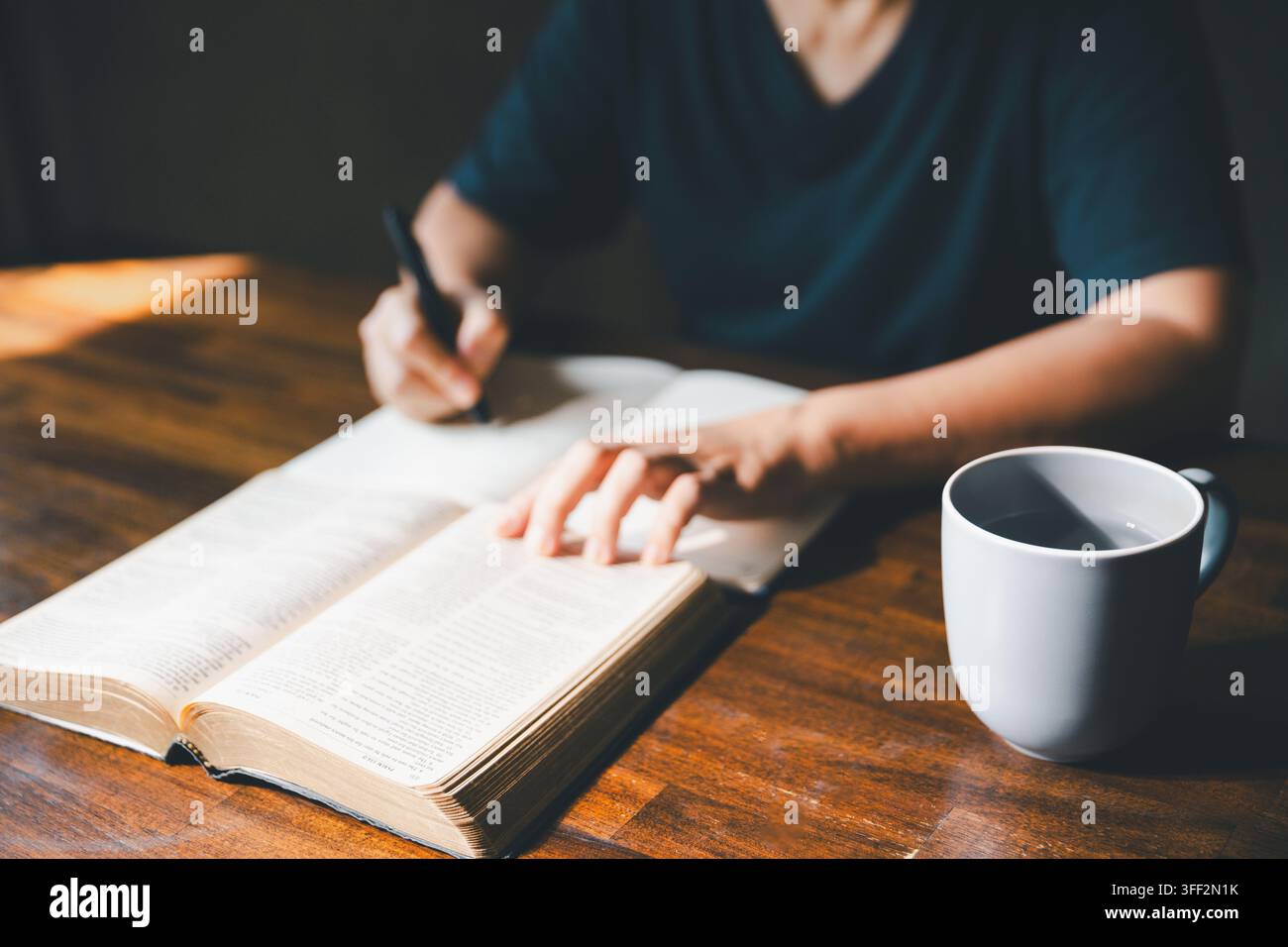Personne adulte écrivant des notes à côté d'une Bible ouverte et d'une tasse sur une table en bois, représentant l'étude des Écritures, la dévotion chrétienne, la réflexion, l'apprentissage de la foi Banque D'Images