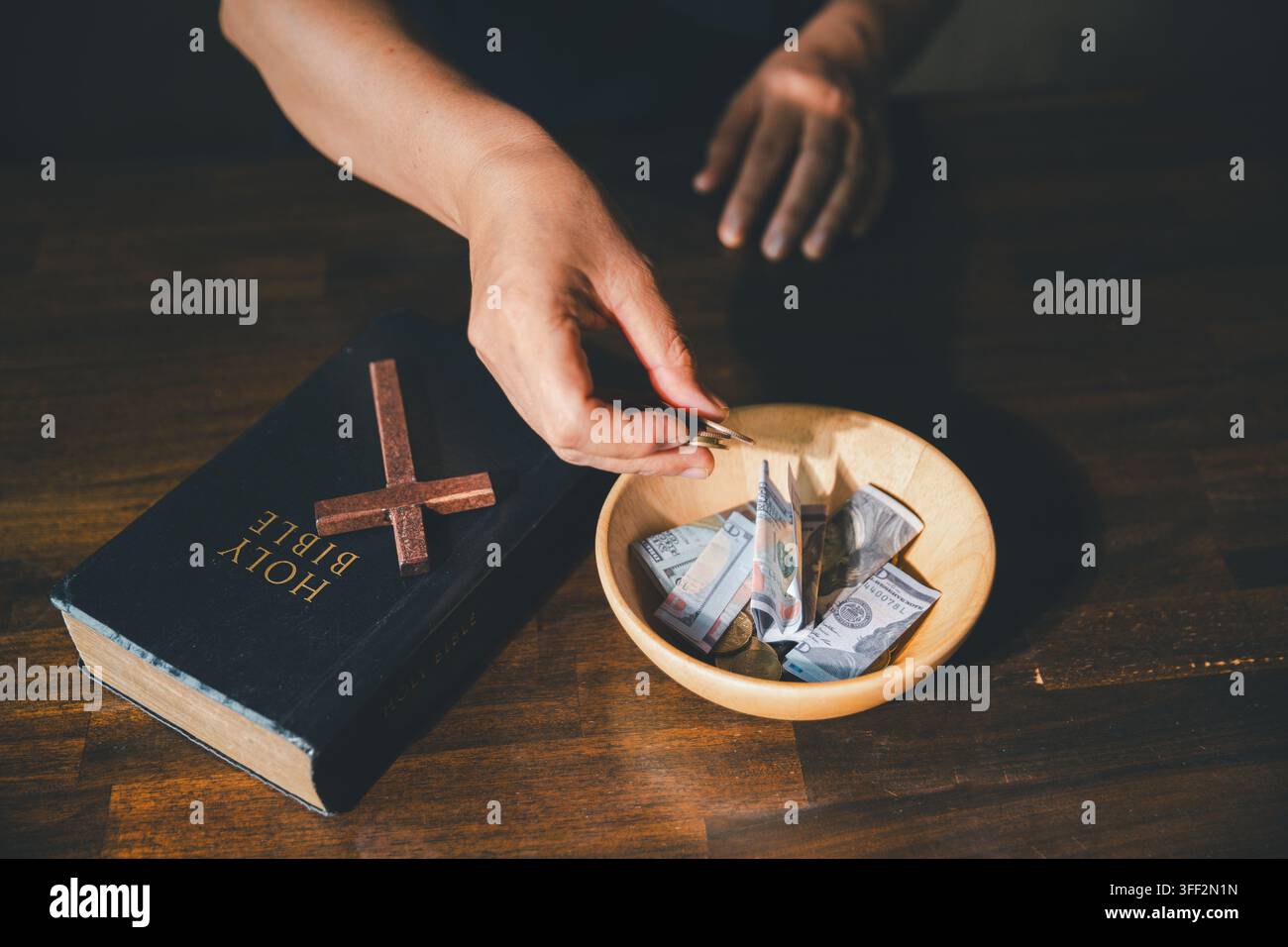 Main plaçant des pièces dans un bol de dons à côté d'une Bible et d'une croix en bois, symbolisant le don chrétien, l'offrande de l'église, la charité, l'intendance, la générosité Banque D'Images