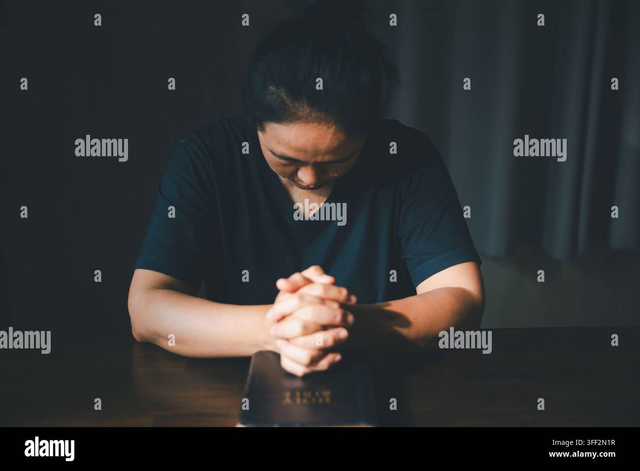Personne adulte priant les mains jointes et la tête inclinée sur une Bible posée sur une table en bois dans une pièce sombre, exprimant la foi chrétienne, la dévotion, l'adoration, h Banque D'Images