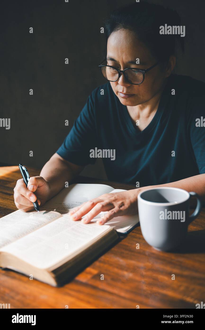 Personne adulte écrivant des notes à côté d'une Bible ouverte et d'une tasse sur une table en bois, représentant l'étude des Écritures, la dévotion chrétienne, la réflexion, l'apprentissage de la foi Banque D'Images