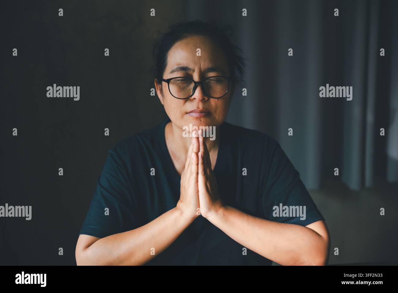 Person adulte portant des lunettes, priant les yeux fermés et les paumes jointes dans une pièce sombre, exprimant la foi, la dévotion, l'adoration, la spiritualité, l'espoir, la paix Banque D'Images